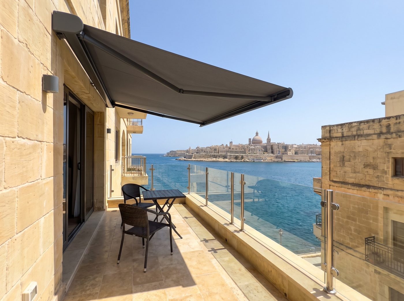 An anthracite cassette awning over a seafront balcony with two chairs, Valletta across the harbour. Illustrative image