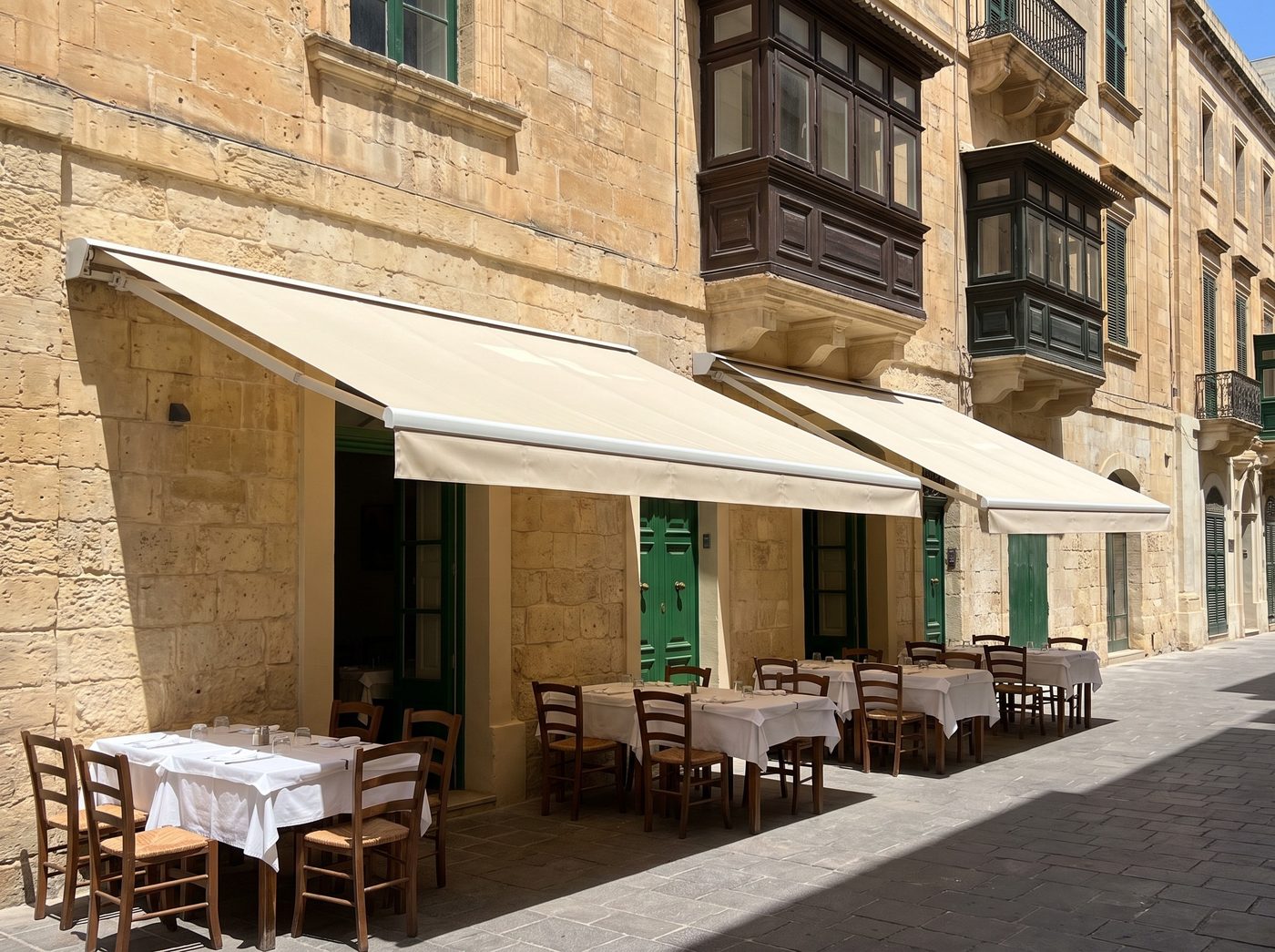 Two cream cassette awnings shading restaurant tables on a limestone street in Valletta. Illustrative image