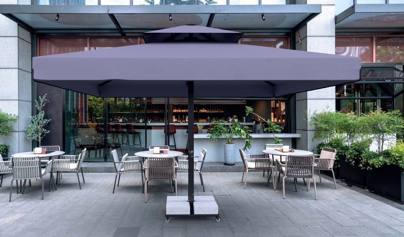 A large square grey parasol on a central pole over round tables on a stone terrace in front of a glass-fronted bar