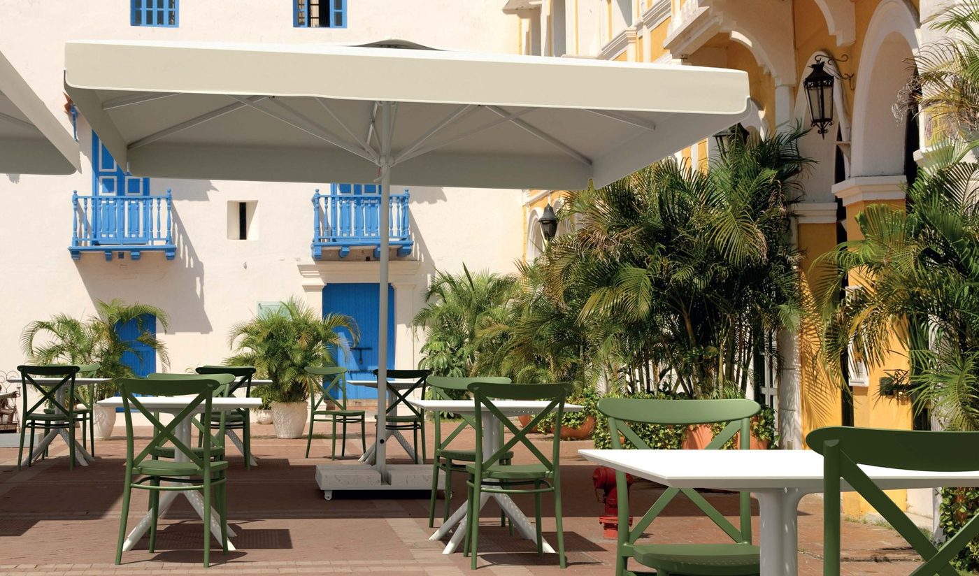 Wide cream parasols over green bistro chairs in a palm-planted courtyard with yellow colonial-style buildings