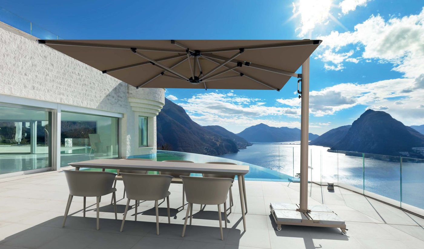 A large taupe side-pole parasol over a dining table on a terrace beside an infinity pool, with lake and mountains beyond