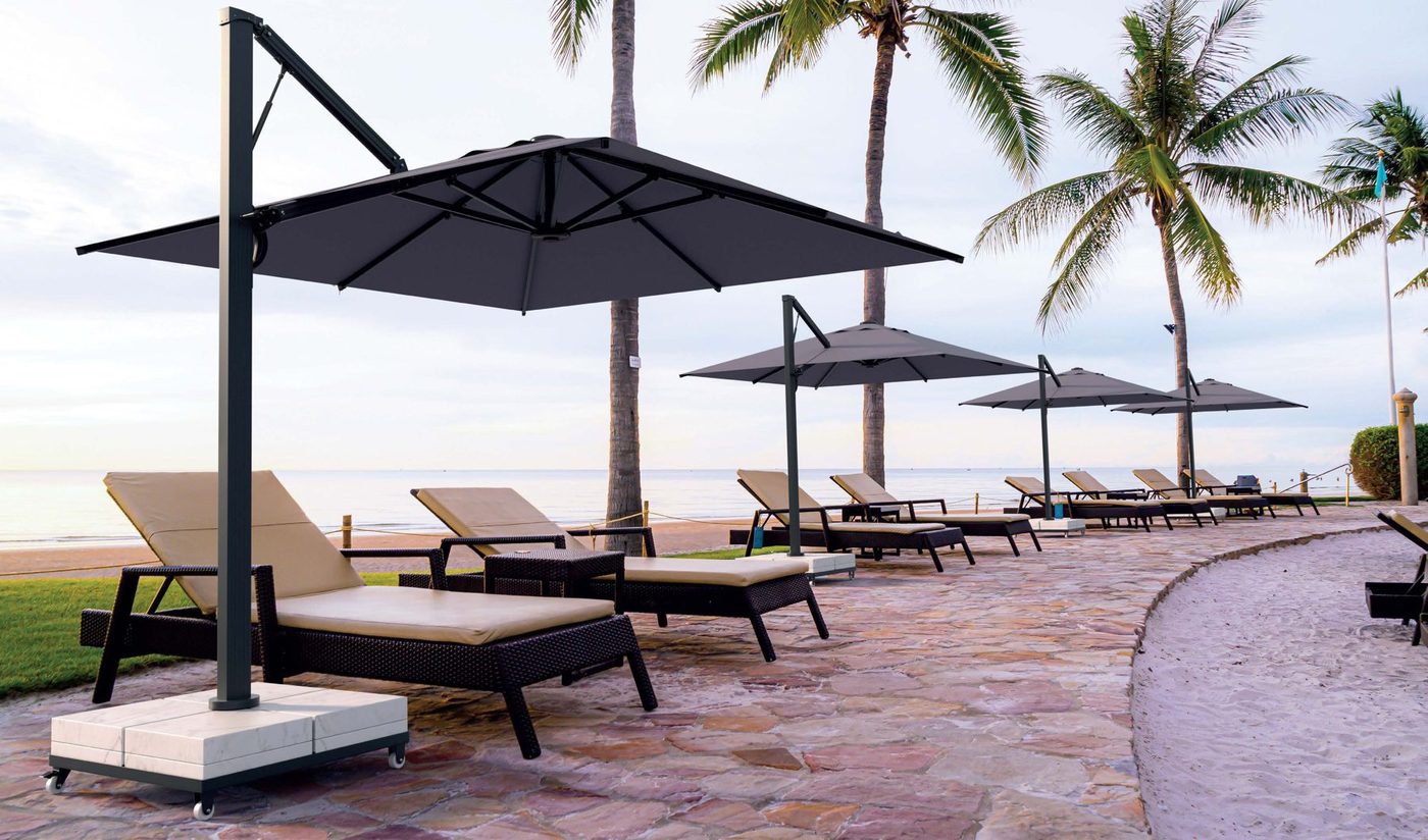 A row of square anthracite side-pole parasols over sun loungers along a palm-lined seafront promenade