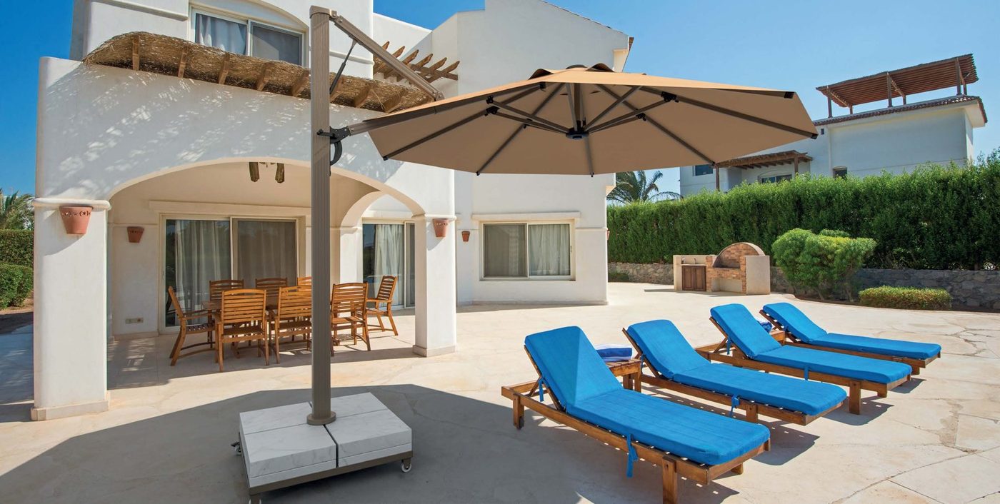 A round taupe side-pole parasol over blue-cushioned sun loungers on a white villa patio