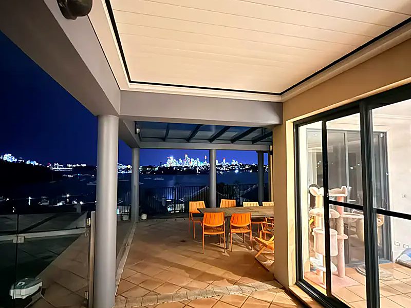 A louvred pergola roof lit from below at night over a tiled terrace with dining chairs and a city view