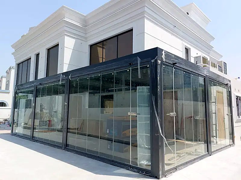 A dark-framed pergola fully enclosed with sliding glass beside a white villa